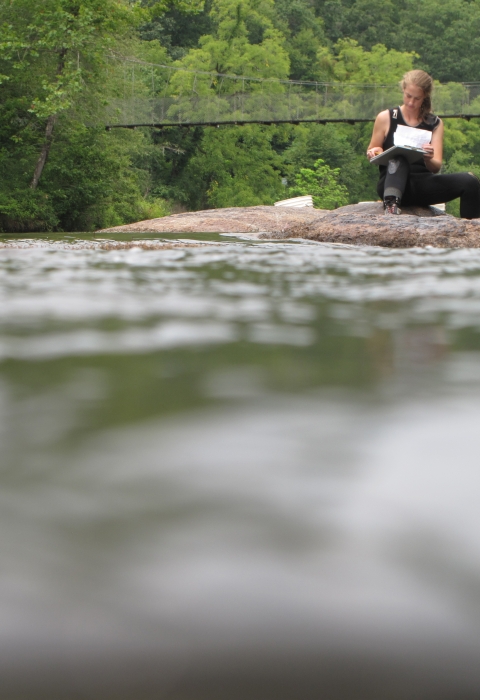 Woman sitting on a rock in a river, writing on a clipboard