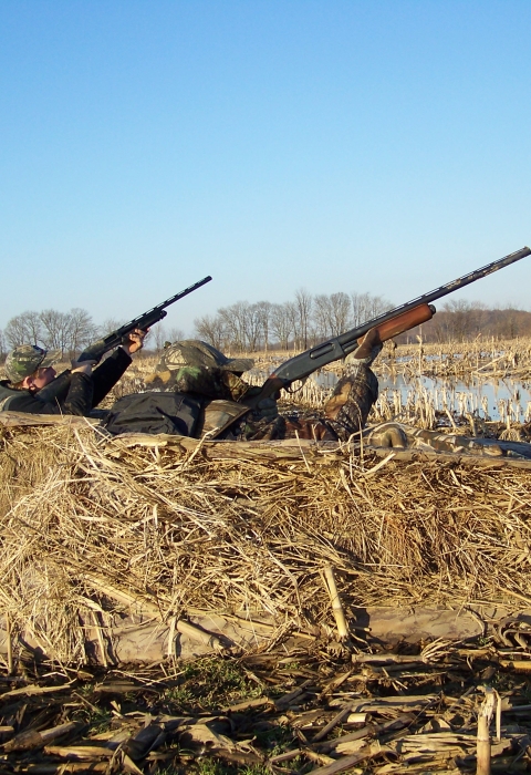 Two hunters in dirt raised camouflaged dirt bunks with shotgun raised. 