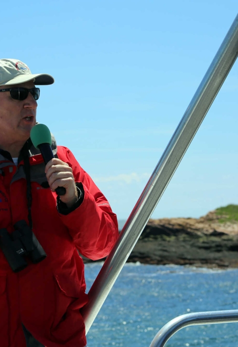 Man stands at the front of a ship holding a microphone. A lihthouse is seen behind 