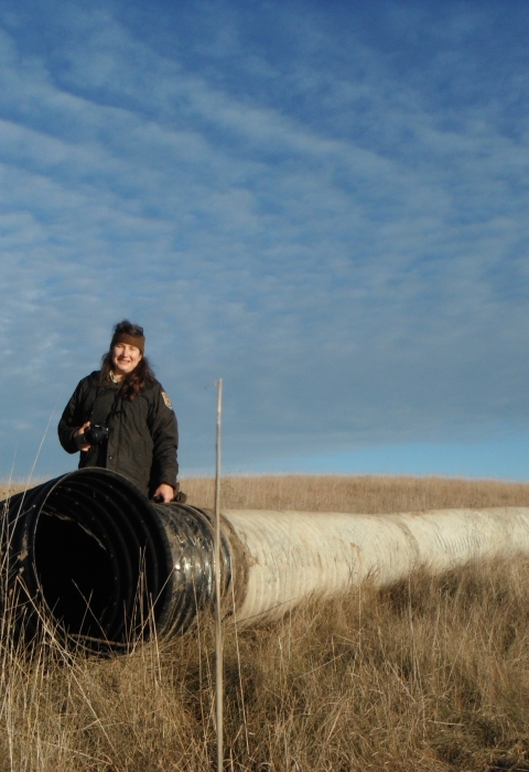 An uniformed adult stands in front of one end of a long drain pipe in a grassy prairie
