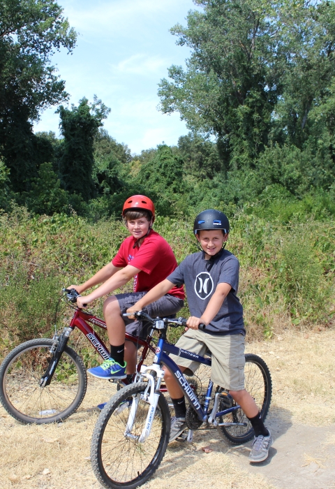 Two children sit on bikes on a trail in front of shrubs and trees