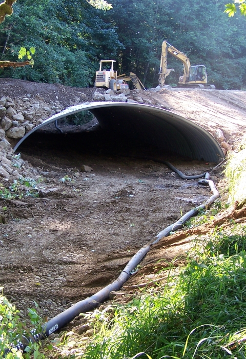 Dry streambed with new culvert for fish passage installed, with heavy machinery on the road above
