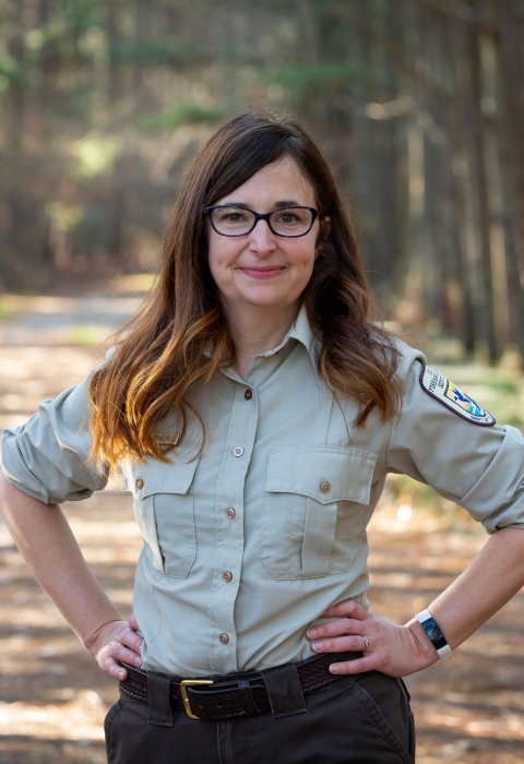 A woman with long brown hair stands on a trail framed by forest. Her hands are on her hips. She wears glasses and a U.S. Fish and Wildlife Service Uniform. 
