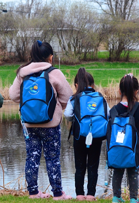 Four kids face towards a pond with backpacks on