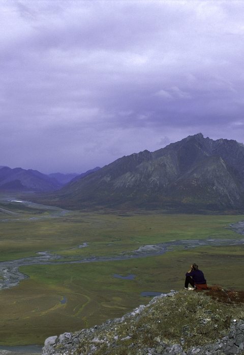 person sitting in front of a stark mountain landscape near a river