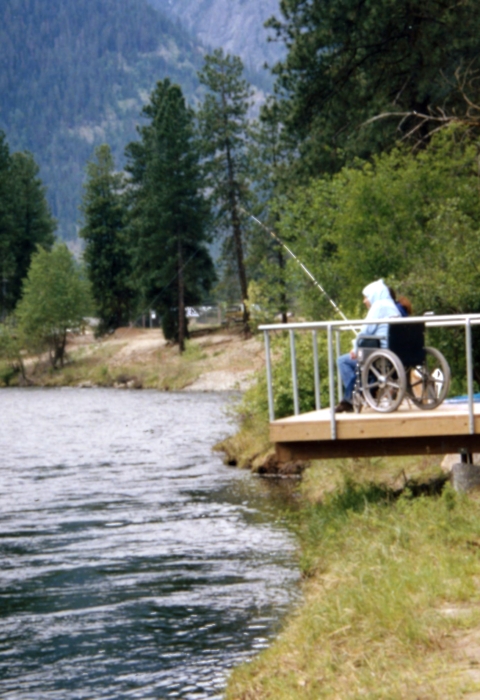 A man wearing a hoodie and sitting in a wheelchair fishes from a platform overhanging a river.