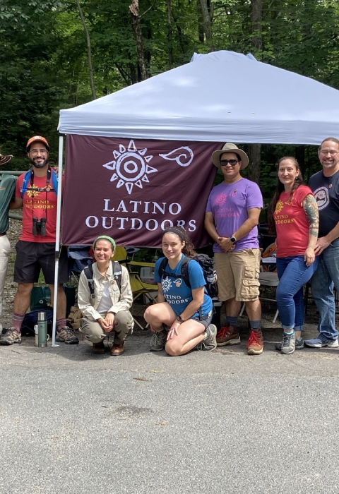 a group of people stand by an outdoor tent. A flag raised on the tent says "latino Outdoors" with a symbol of a sun above it. THe people are smiling wearing hats, binoculars and backpacks