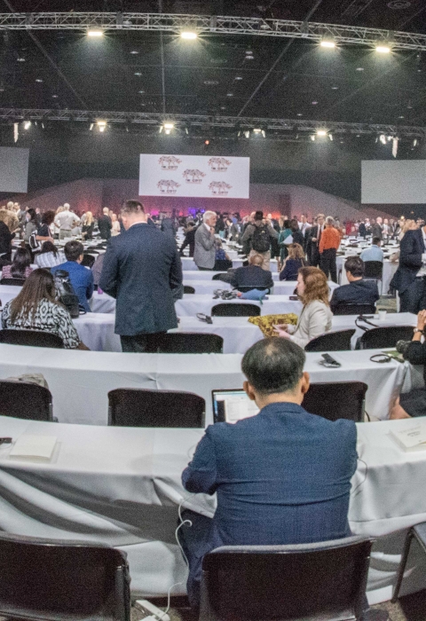 A fish-eye shot of an international convention, many tables, flags, and people having conversations, indoors.