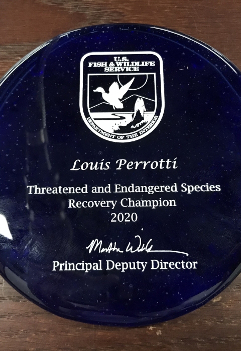 Round blue glass award featuring a U.S. Fish and Wildlife Service logo. Text reads: Louis Perrotti Threatened and Endangered Species Champion 2020 and is signed by Principal Deputy Director Martha Williams