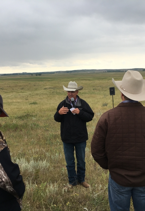 A man wearing a cowboy hat speaks to a small group of people on a grassland