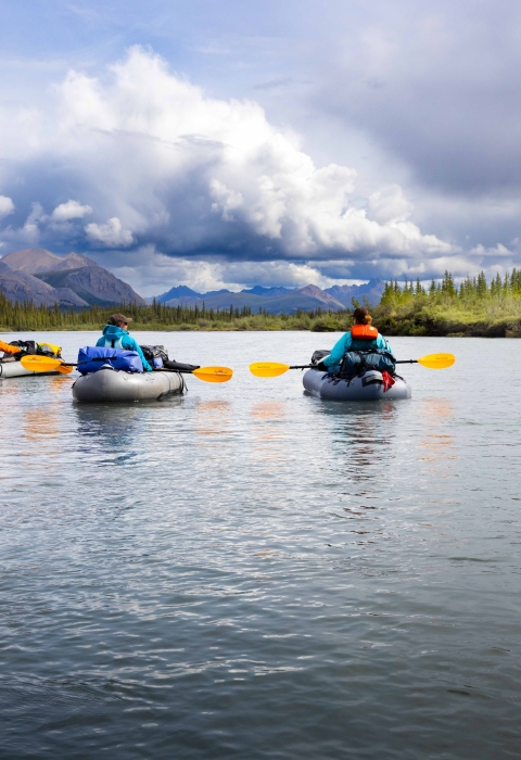View of four people floating a river with mountains and a cloudy sky in the background. 