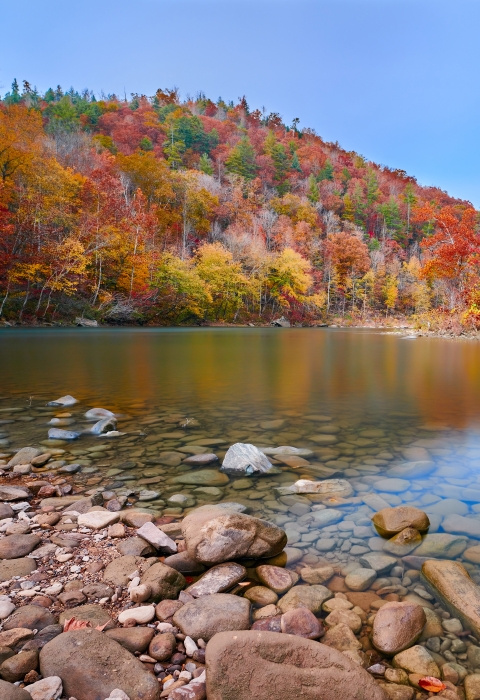 A lovely river with a rock bank on both sides. In the background the trees are various vibrant fall colors. 