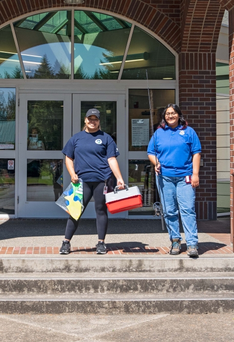 Two Service interns posing with fishing gear in front of a library. 