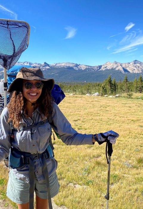 A woman smiles for the camera while holding hiking poles and while wearing a backpack with an attached net on her back
