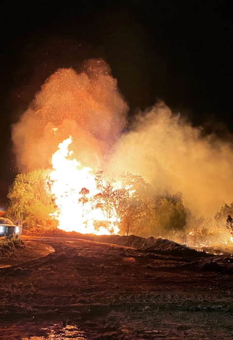 A fire burns in the night. There is a firefighter standing to the right of the burn.