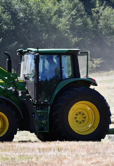 A green and yellow tractor tows a mowing attachment across a dusty field of grass with evergreens in the background.