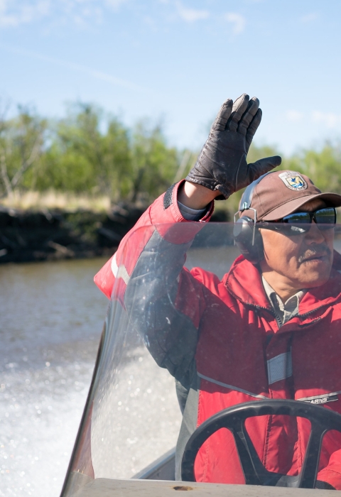 Close up of Chris Tulik operating a boat on a river while waving his hand at someone outside of the image. 