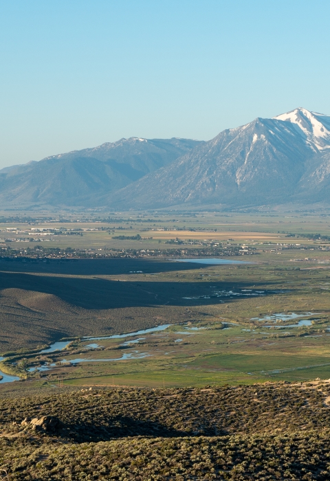 A view looking into Carson Valley during sunrise. Tall mountains are off in the distance with a valley filled with sagebrush below. A river cuts through the valley.