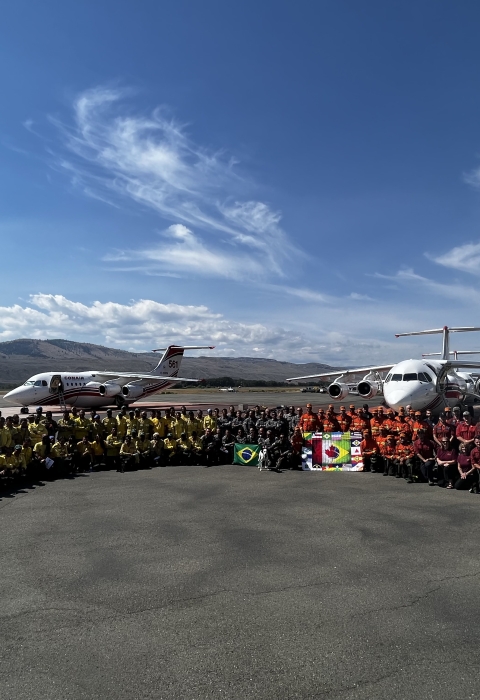 A group of people stand together in a semi circle on a tarmac in front of aircraft used for firefighting efforts. The group has flags out and are all in firefighter clothing or agency t-shirts.