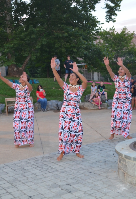  7 people in similar dresses with hands raised to left in unison dance outdoor amphitheater 