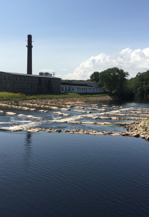 A river with rocks placed in lines flows by an old brick mill complex