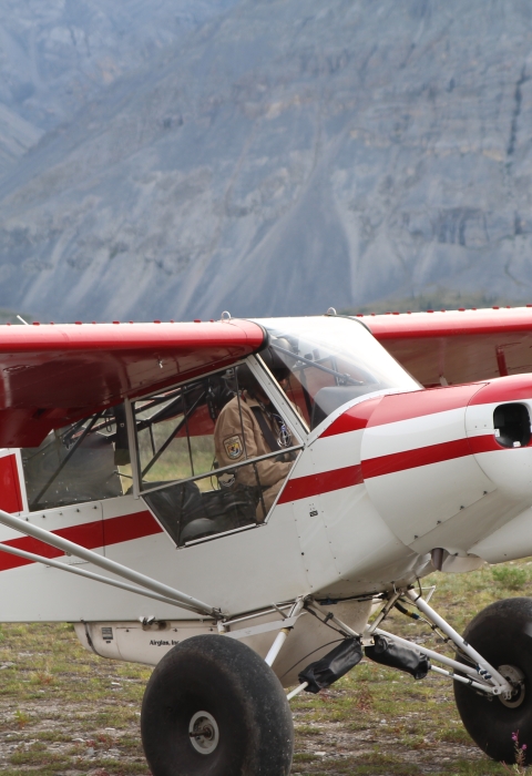 Close up of a Refuge Federal Wildlife Officer in a Top Cub bush plane as he prepares for take off from a dirt airstrip with mountains covering the background just out of focus. 