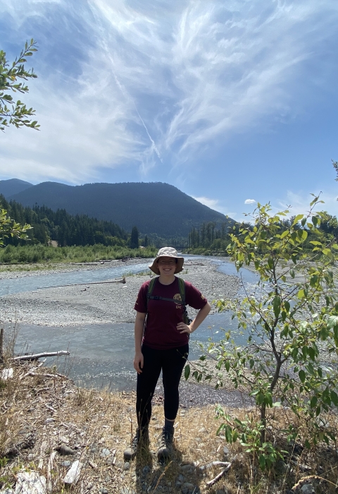 Service intern, Kathleen Gerard, poses in front of the Elwha River with mountains in the background.