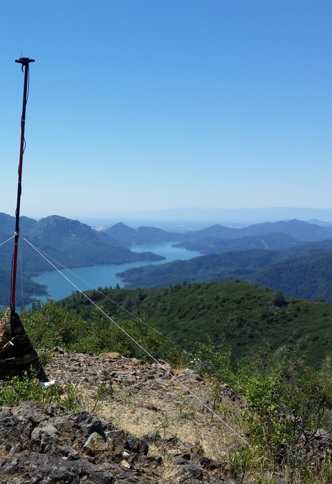 A tall pole on the top of a mountain with a microphone at the top. This pole is secured with anchors to the rocks below. In the background is a bright blue river flowing through the valley of mountains. 