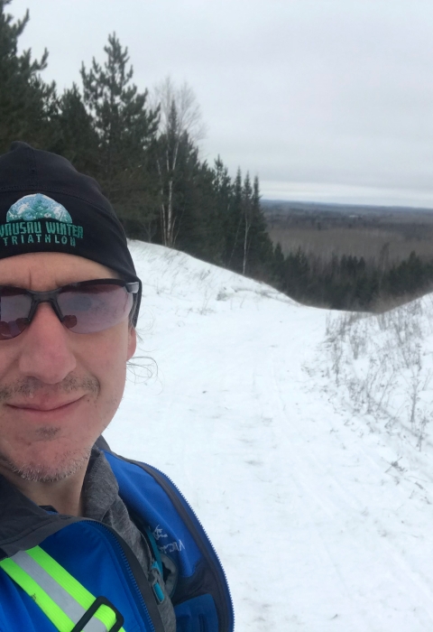 A man in a knit hat, sunglasses and a blue jacket smiles at the top of a snowy trail.