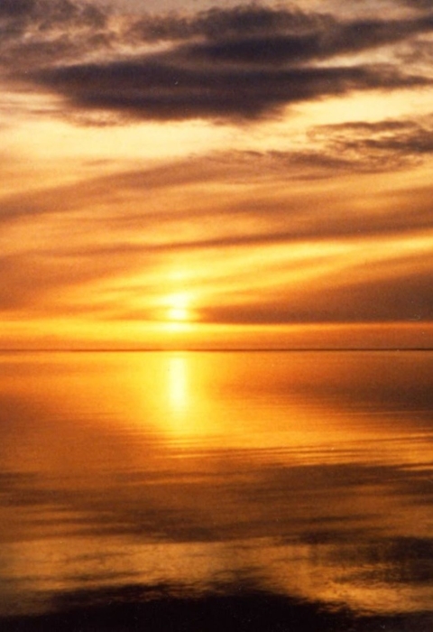 An orange sunset over calm water. The sky is scattered with clouds and the water is reflecting the sun and sky.