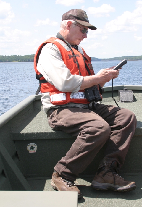 A man in an orange vest and refuge clothing sits on the bow of a boat in the middle of a lake looking at surveying equipment