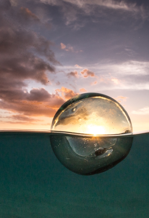 A glass ball floats in the ocean with the sun setting through it.