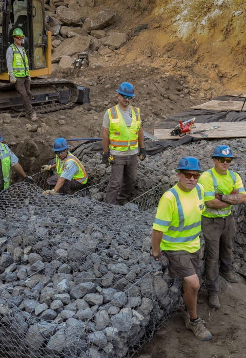 Ten people wearing bring yellow shirts and blue hard hats pose near a newly constructed rock wall