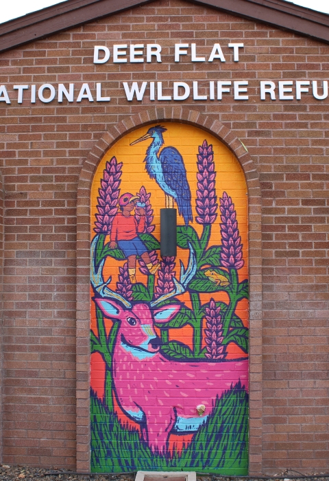 Colorful mural of a deer, boy and heron on a brick building that reads "Deer Flat National Wildlife Refuge"
