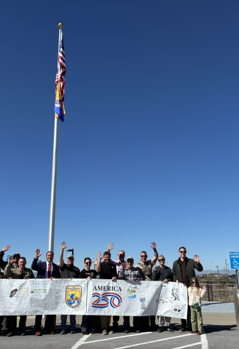 Group of people standing outdoors holding a USFWS-America 250 banner beneath a flagpole; flags flat