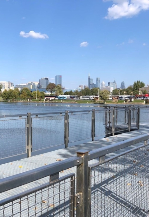 A view of a new pier with metal railings and synthetic decking jutting into a river with a city skyline in the distance