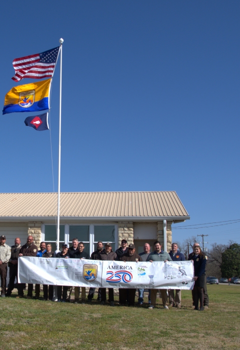 People hold FWS A250 banner at base of flagpole with US flag, FWS flag, fish pennant flying