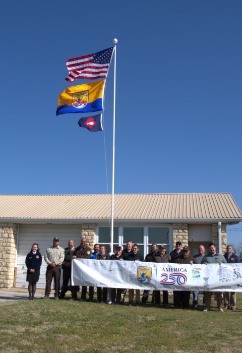 People hold FWS A250 banner at base of flagpole with US flag, FWS flag, fish pennant flying