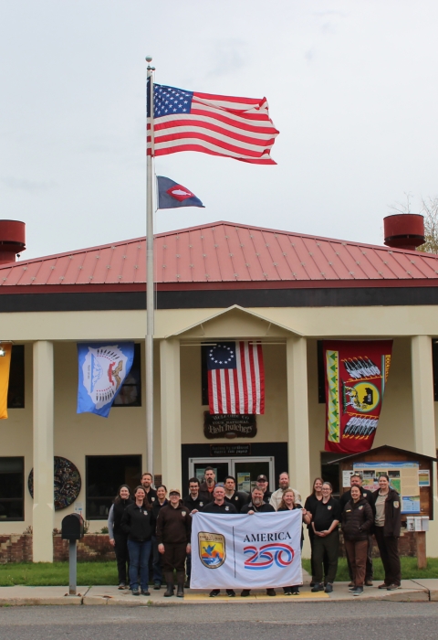 A group of people in brown holding a white “America 250” flag stand in front of a tan building with a red roof. Flags from USFWS, the Confederated Tribes and Bands of the Yakama Nation, the Betsy Ross flag, the Confederated Tribes of the Colville Reservation, and the Bureau of Reclamation flag fly behind. US flag and fish pennant fly above