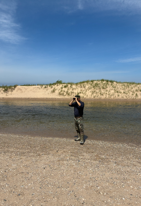 A man standing on a beach looking through binoculars away from the water.