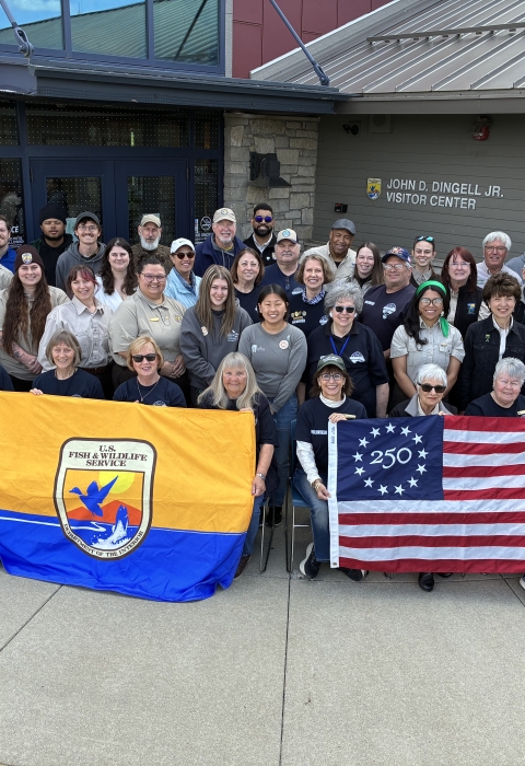 Large group posing outside a building with sign on it saying “JOHN D. DINGELL JR. VISITOR CENTER.” They’re holding USFWS flag and Freedom 250 U.S. flag; blue mascot stands at right