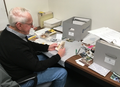 A man sits at a desk holding some photographs. Spread out in front of him are archival folders, boxes and more photos that he is sorting to be processed and put into archival storage.