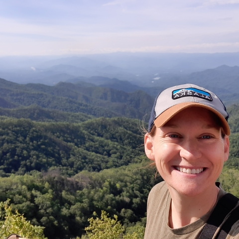 A caucasian woman wearing a baseball cap and brown shirt, in front of a wooded hilly overlook