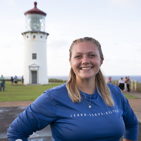 A blond woman in her 20's in a blue uniform stands in front of a lighthouse. She is smiling. 