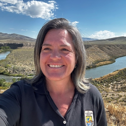 Selfie of Sue Kerver near a river bend with a low brush landscape and spotted clouds in a bright sunny sky. 