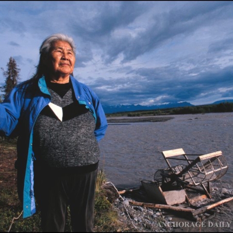 An elder woman stands on the bank of a river with a fish wheel on the gravel shore. She holds a walking stick and looks in the distance beyond the camera.