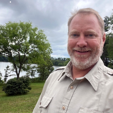 Refuge employee in a tan uniform smiling with tree and water in background