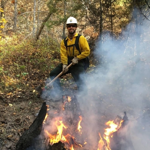 Chris in a hard hat and protective gear holding an ax near a fire