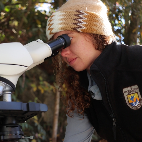 Madison wearing a tan toboggan and a black jacket with USFWS emblem looking through binoculars