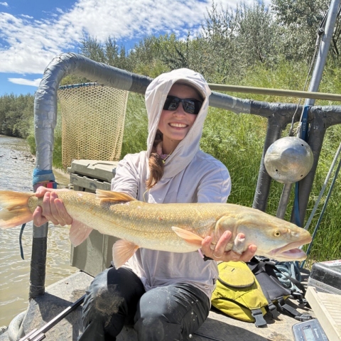 a woman in a gray hoodie and sun glasses smiles while sitting in a boat holding a large fish
